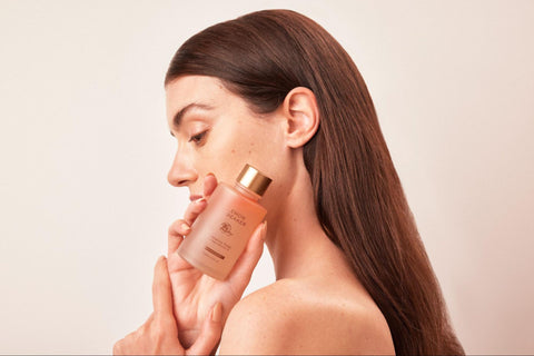 A woman with clear and glowing skin holds up a bottle of hydrating toner.