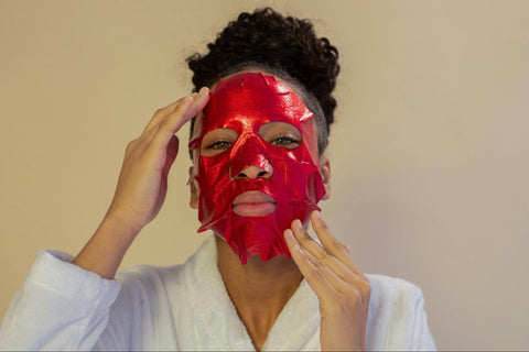 A woman with her hair pulled back applies a red-colored sheet mask.