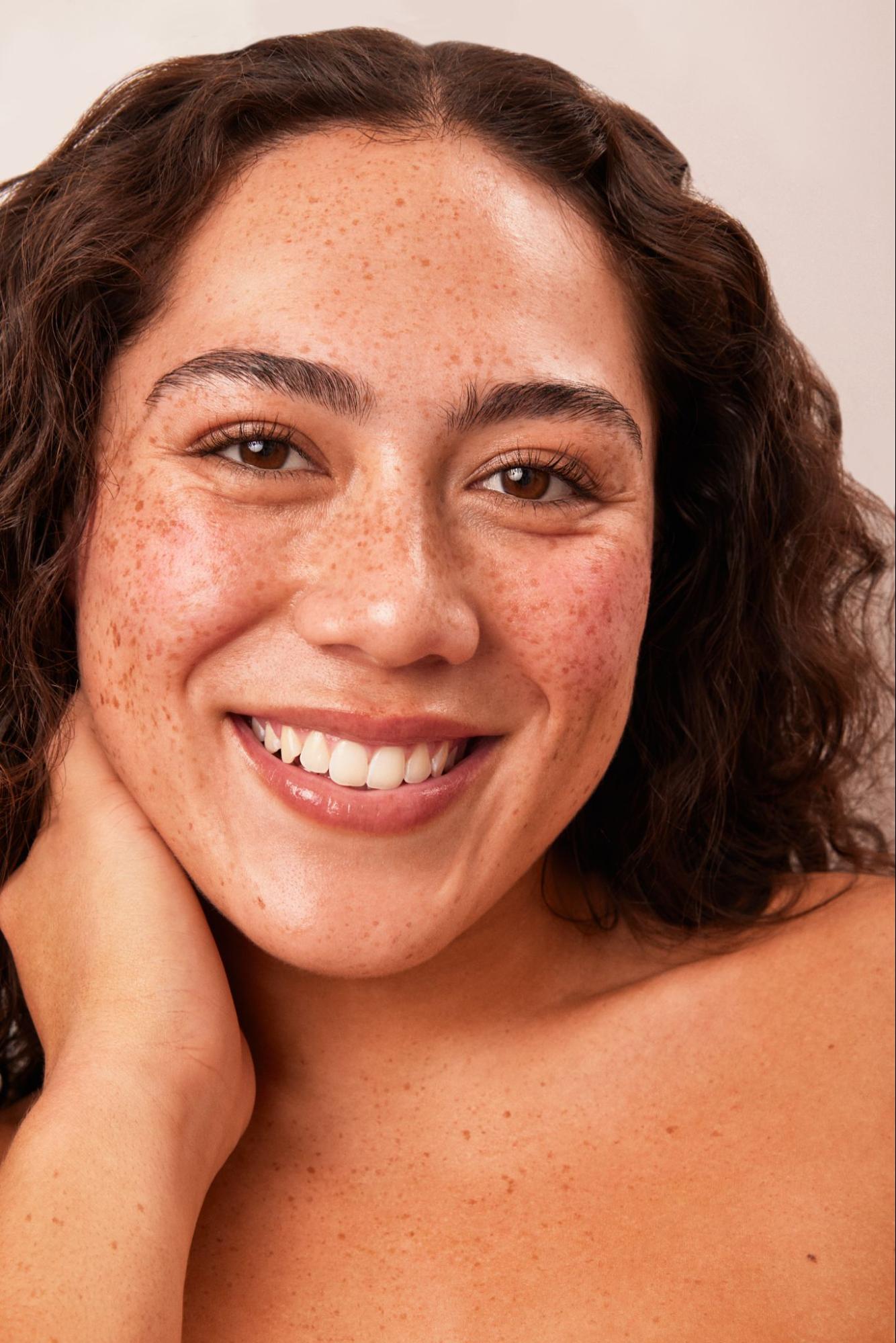 A woman with glowing, radiant skin smiles while resting her face on her hand