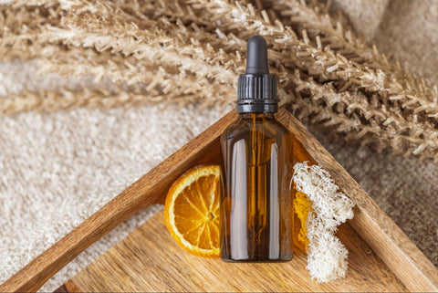 An amber bottle of serum lays atop a dried orange slice and a bit of loofah in a wooden tray.